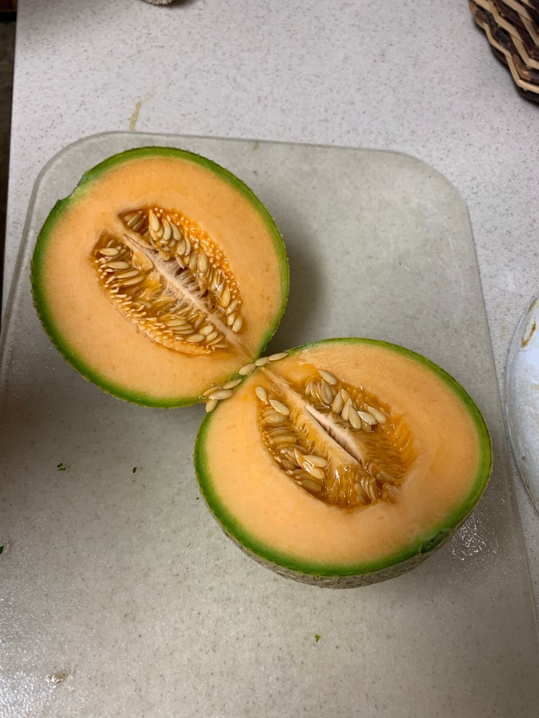 A ripe cantaloupe cut in half with seeds showing resting on a cutting board on a counter.
