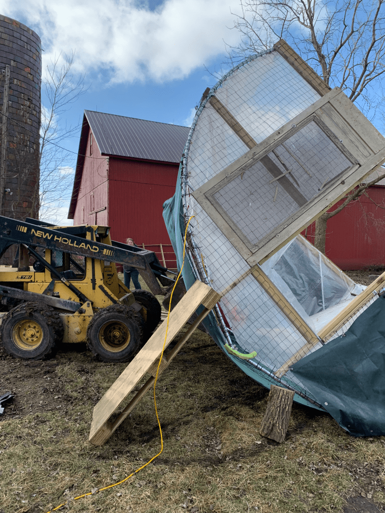A pickture of the upside down coop with a skidsteer. The coop is propped on a log, a pallet, and the bucket of the skidsteer.